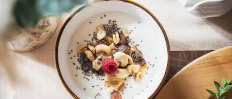 A bowl of yogurt topped with seeds, nuts, and a raspberry, placed on a table with a cup and a plant in the background.