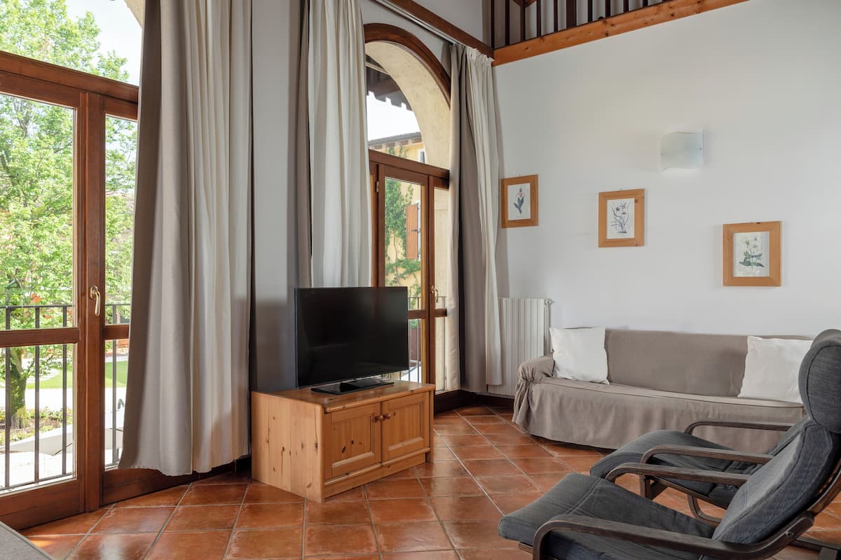 A living room with a television on a wooden cabinet, a sofa, and a chair. There are large windows with curtains, a view of greenery outside, and a tiled floor.