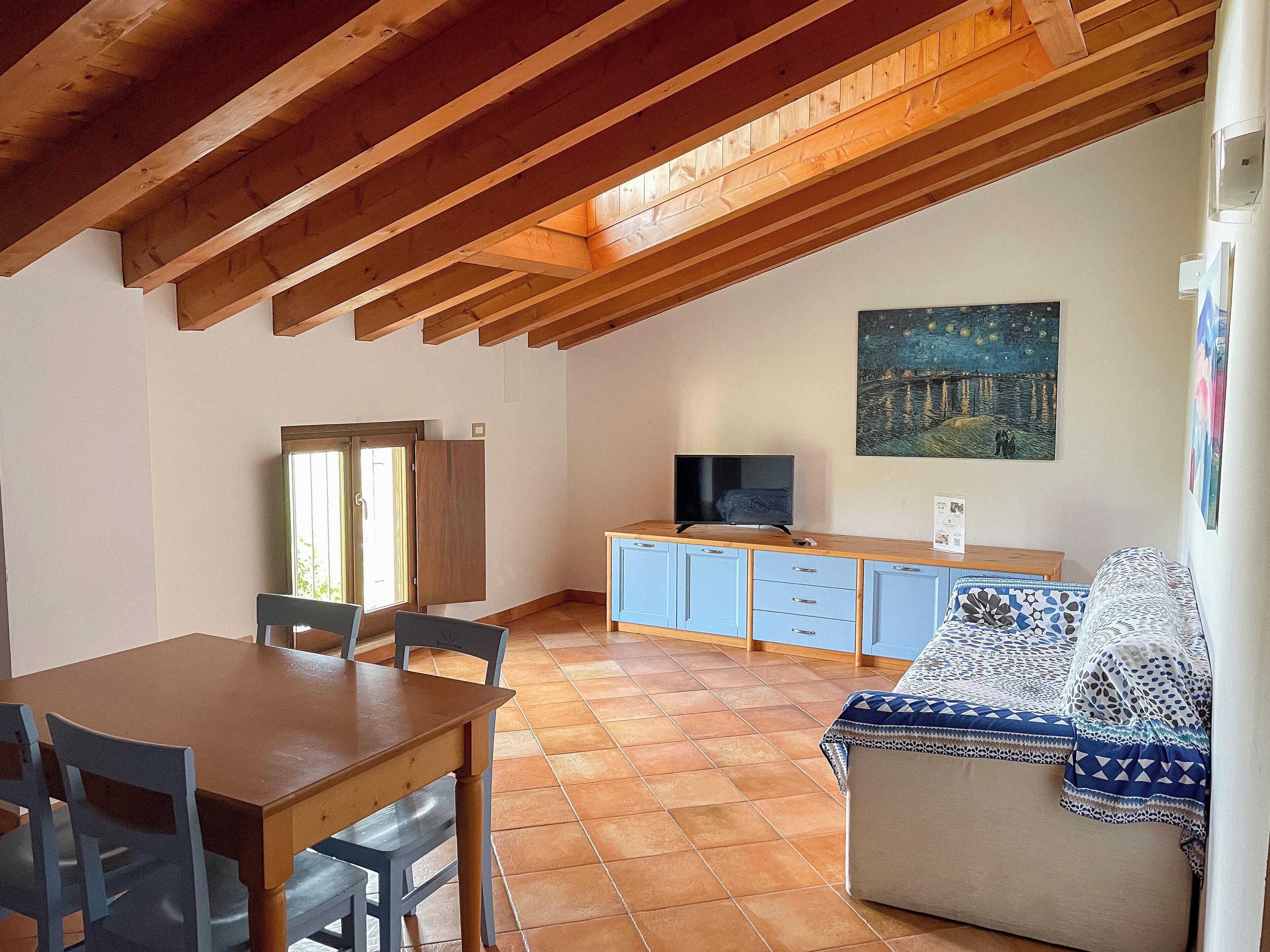 A dining area with a wooden table and chairs, a TV on a wooden cabinet, and a painting on the wall.