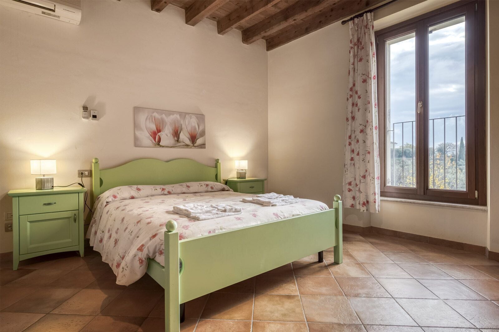 A bedroom with a green bed, two bedside tables, a nightstand, a window with curtains, and a painting on the wall.