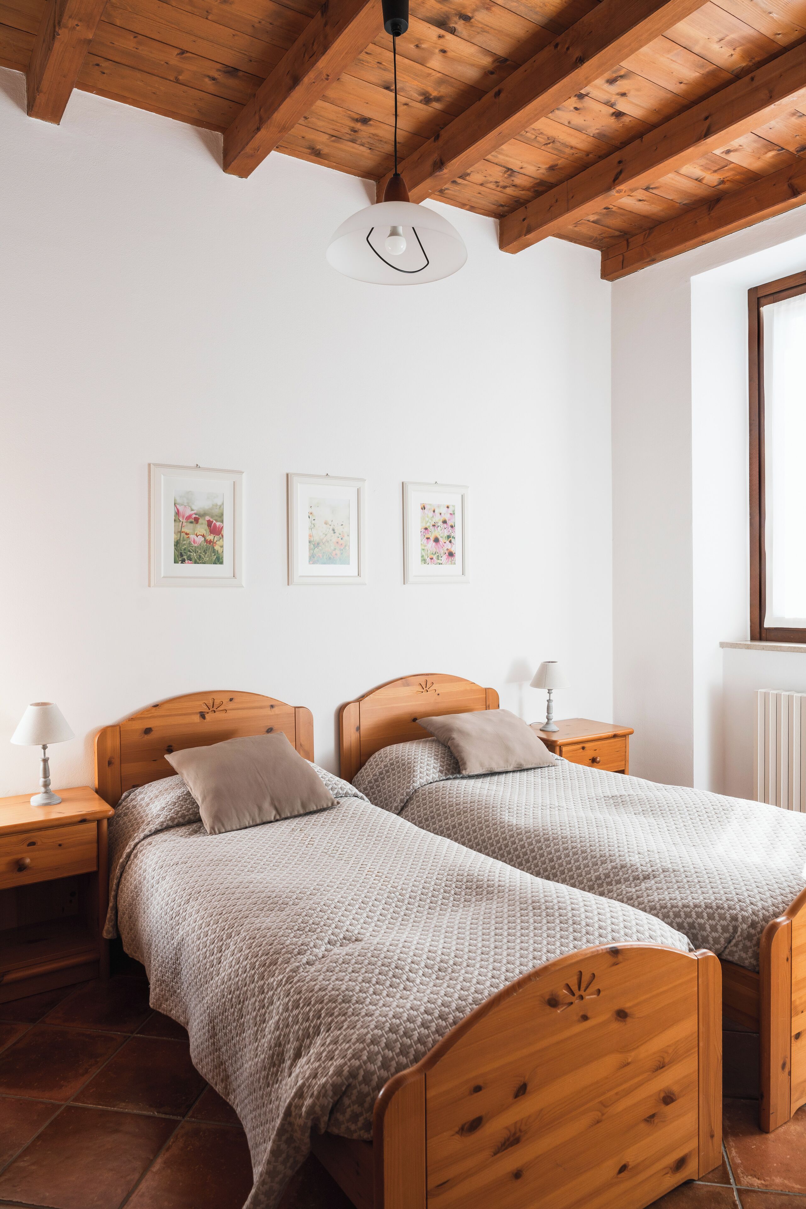 A bedroom with two wooden beds, white walls, and a wooden ceiling. There are framed pictures on the wall, a window, and a radiator.
