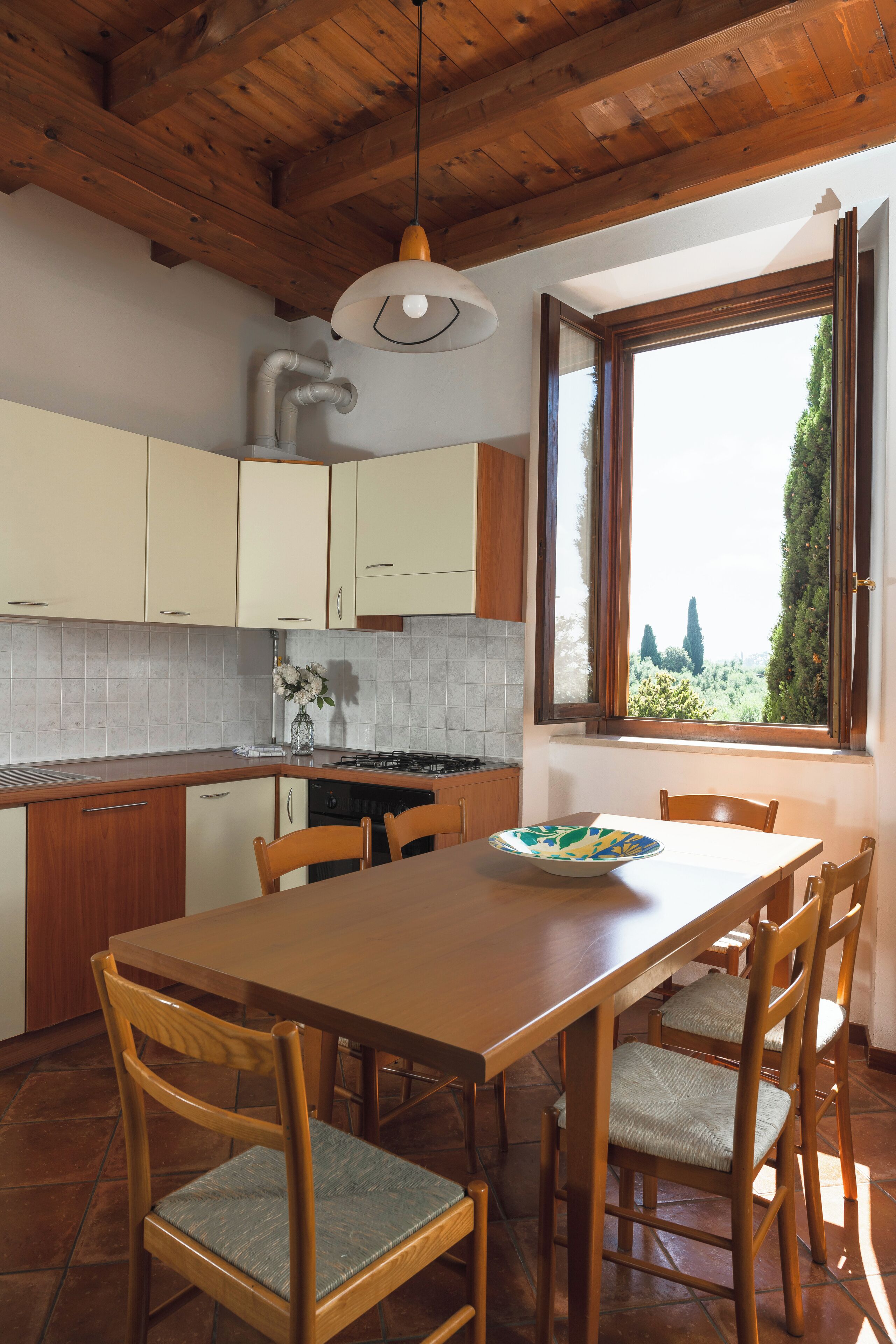 A kitchen with wooden cabinets, a dining table with chairs, a window with a view of trees, and a hanging light fixture.