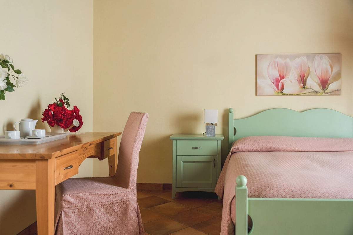 A room with a wooden desk, a chair, a nightstand, a bed with a green headboard, and a wall-mounted painting of flowers.