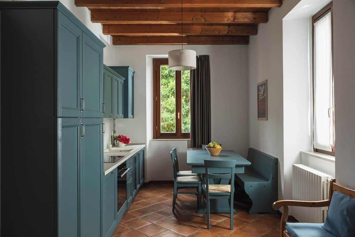 A compact kitchen with teal cabinetry, a dining area with a table and chairs, a window with a view of greenery, and a hanging light fixture.