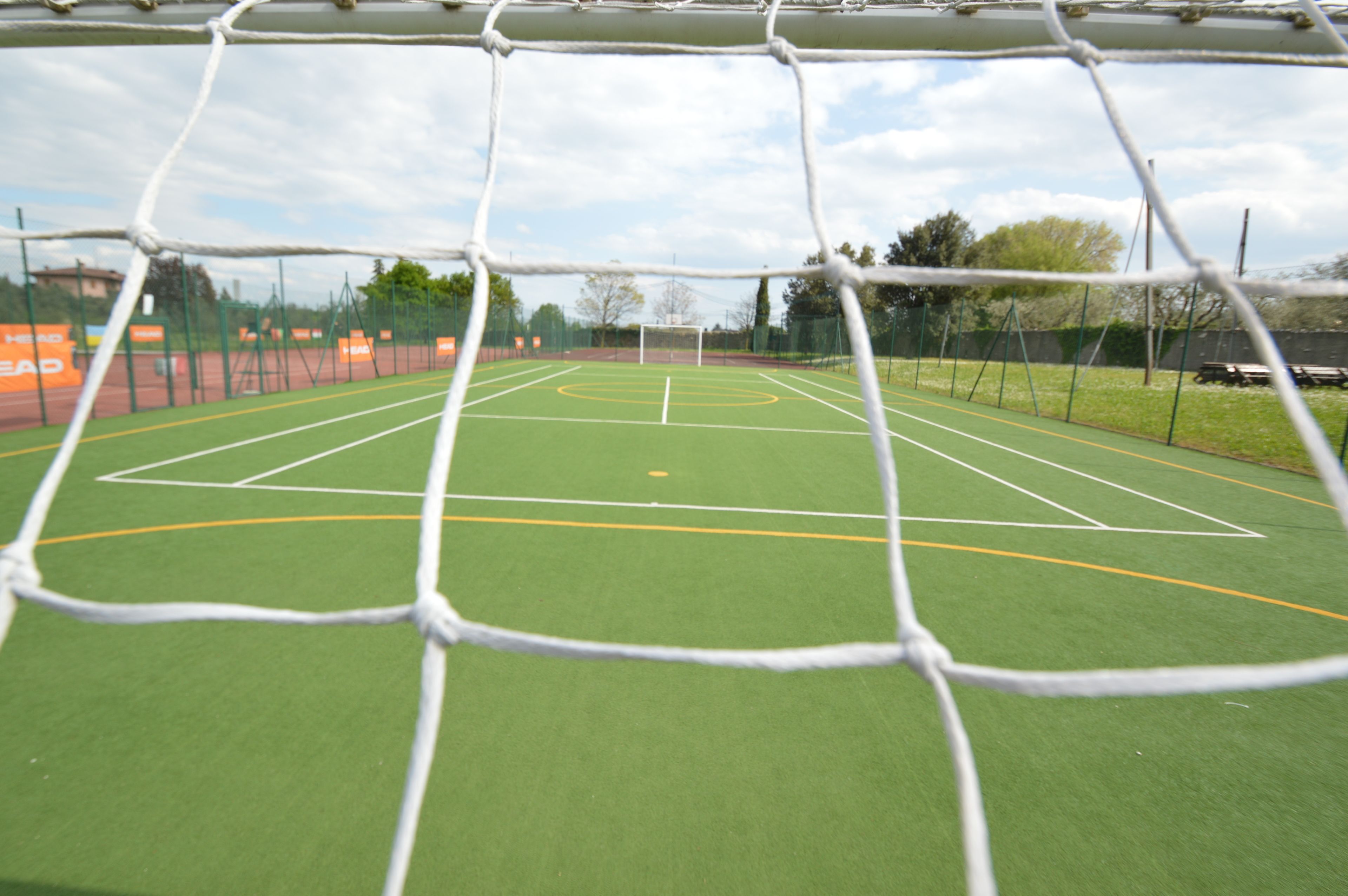 A view through a soccer goal net, revealing a green artificial turf field with white and yellow markings, surrounded by a fence and trees in the background.