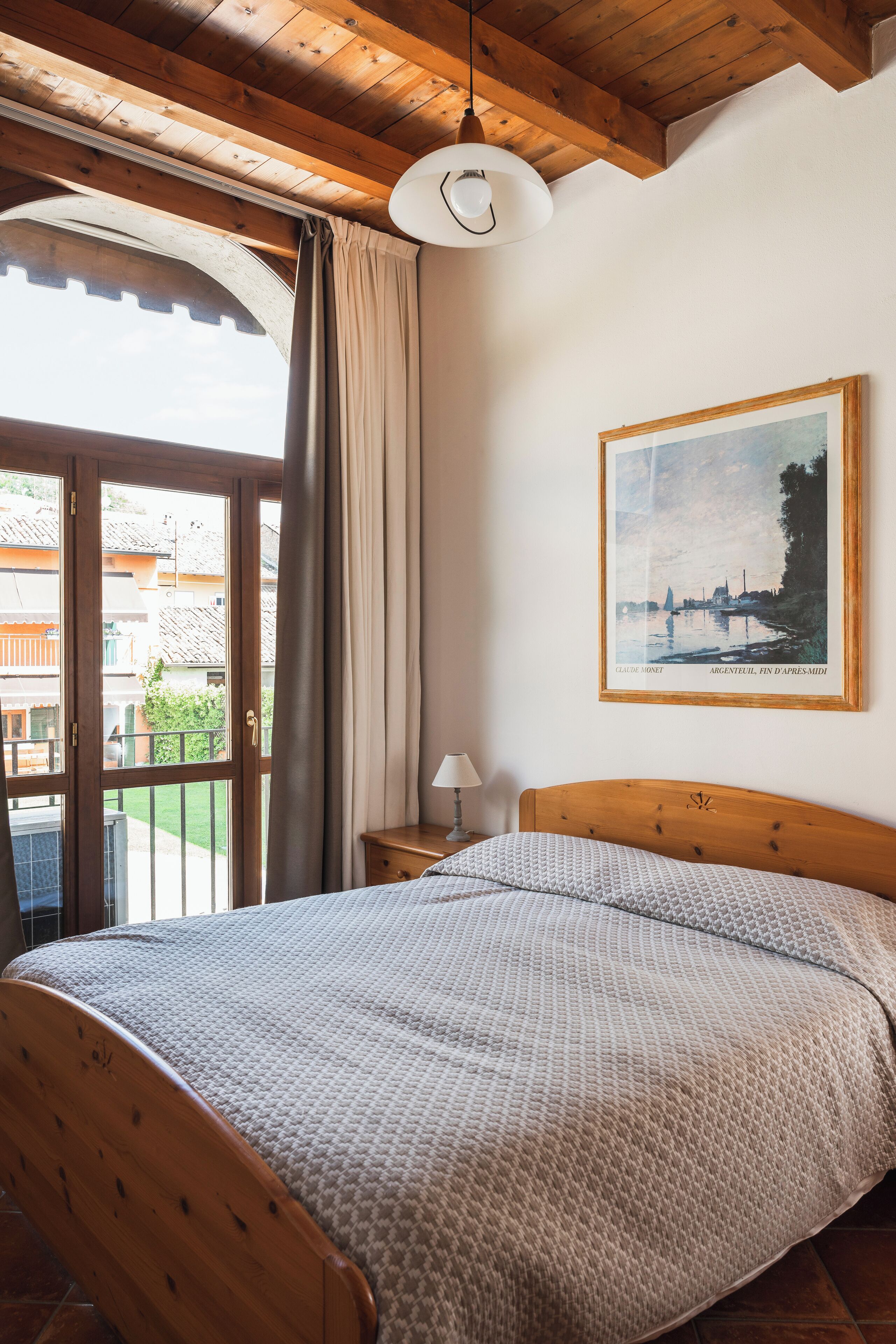 A bedroom with a wooden bed, a nightstand, a lamp, a painting, and a view of the outside.