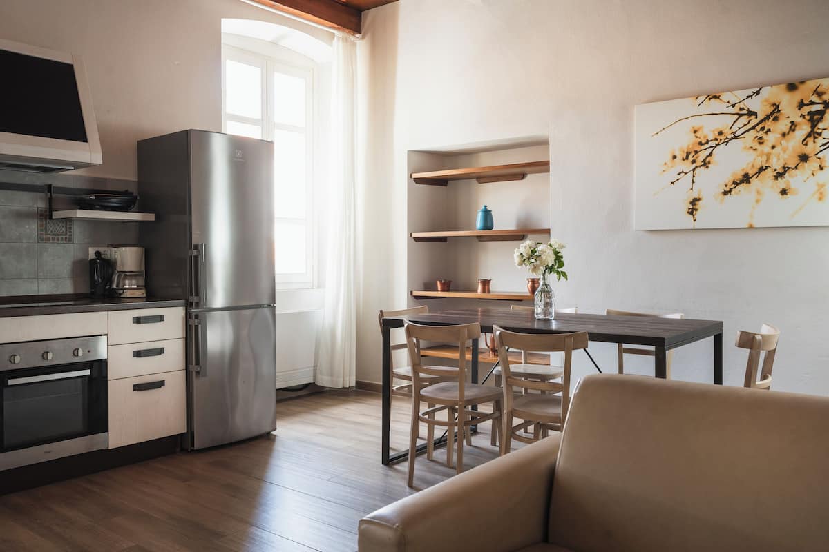 A modern kitchen with a built-in oven, a large refrigerator, and wooden flooring. There is a dining area with a table and chairs, and a sofa in the foreground.