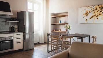 Cocina moderna con horno empotrado, un refrigerador grande y piso de madera. Hay un comedor con una mesa y sillas, y un sofá al frente.