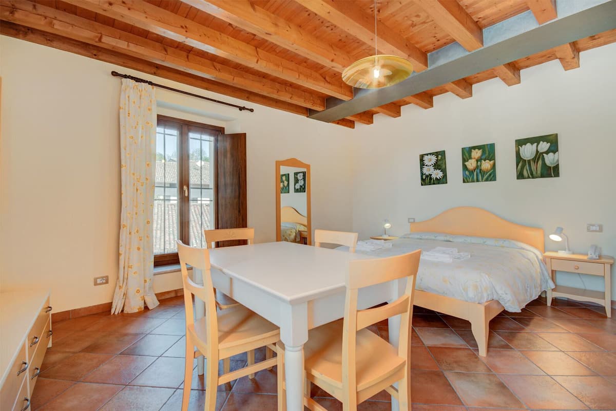 A bedroom with a wooden ceiling, a bed with a headboard, a bedside table with a lamp, a window with curtains, and a dining table with chairs.