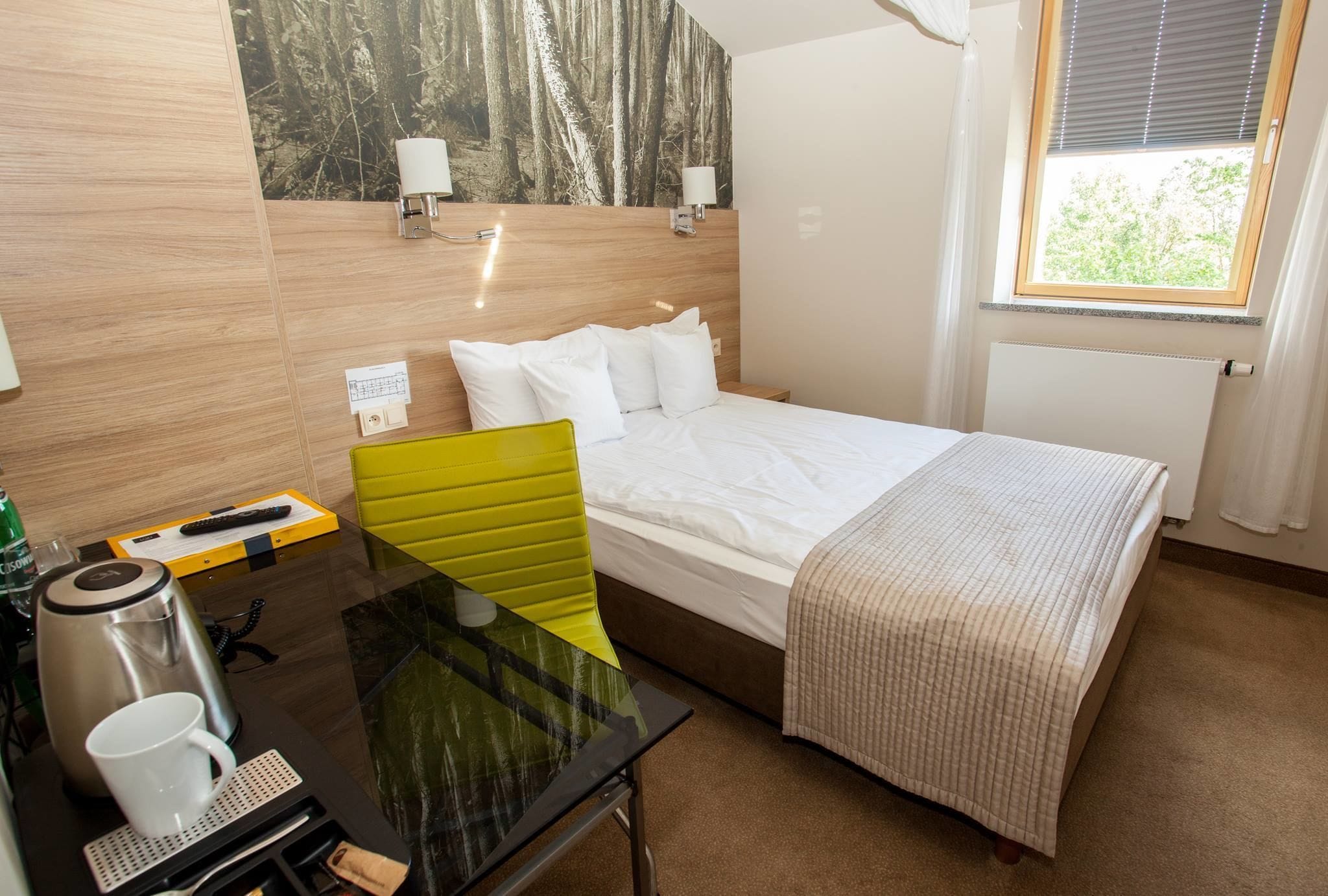 A hotel room with a bed, a desk with a coffee maker, a chair, and a window with blinds.