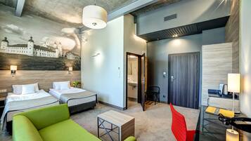 Une chambre d’hôtel avec deux lits, un canapé vert, un bureau avec une lampe et une salle de bain visible à travers une porte ouverte.