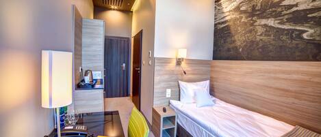 A hotel room with a bed, bedside table, lamp, chair, desk with a lamp, and a painting on the wall.
