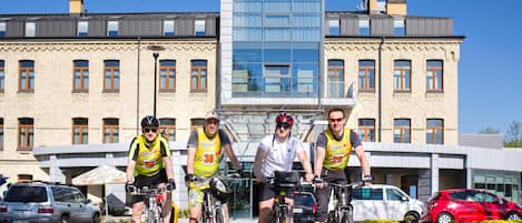 Un hotel de fachada moderna, cielo despejado y cuatro ciclistas adelante.