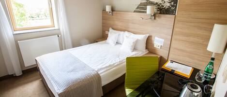 A hotel room with a bed, a green chair, a small table with a kettle, a bottle of water, and a lamp.