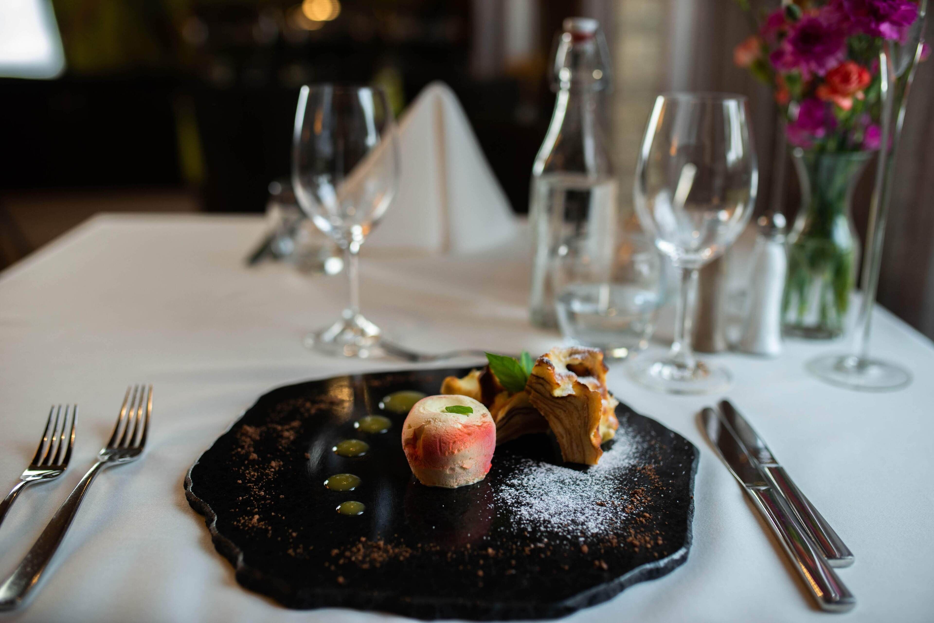 A dessert plate with a scoop of ice cream, a piece of cake, and a mint leaf on a black plate, accompanied by a fork and knife on a white tablecloth.
