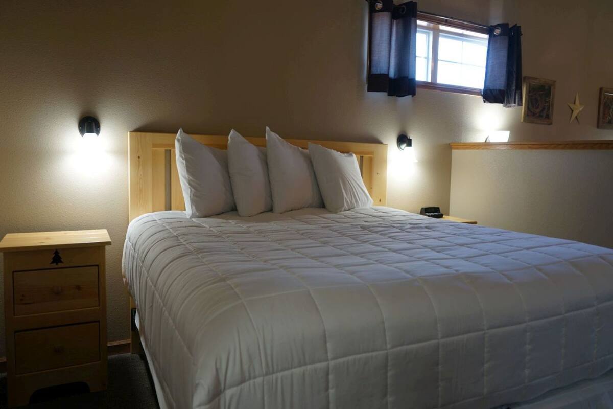 Cama bem arrumada com cabeceira de madeira, mesa de cabeceira com gavetas e luminária fixada na parede.