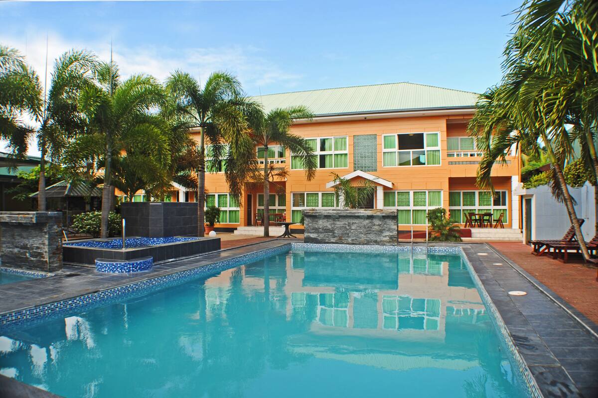 Una piscina con borde de azulejos, rodeada de palmeras y un edificio con múltiples ventanas y techo de tejas.