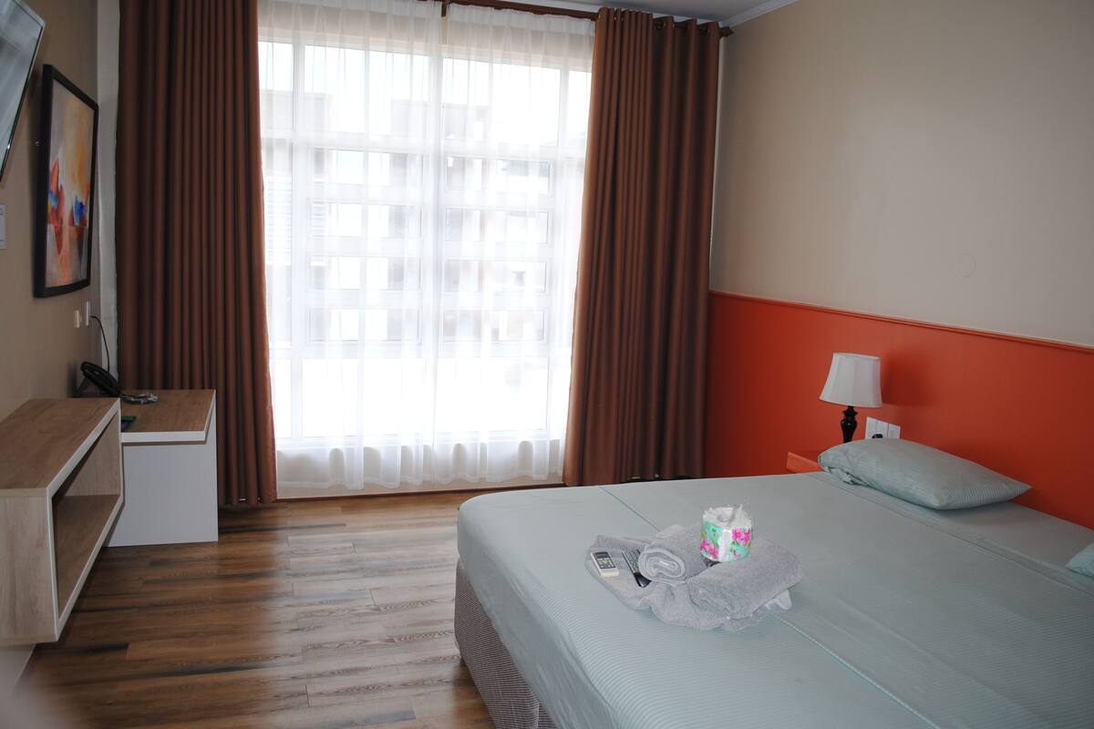 Una habitación de hotel con cama, escritorio, silla, lámpara y ventana con cortinas.