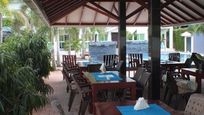 Un área de comedor al aire libre con mesas y sillas, una sección cubierta y una piscina al fondo.
