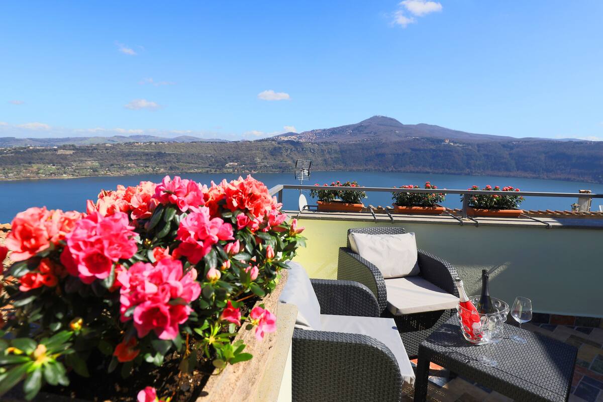 A terrace with wicker furniture, a bottle of champagne, and a glass on a table, overlooking a lake with mountains in the background.