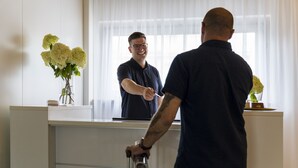 A reception area with a counter, a vase of flowers, and a window with curtains. -  2Places Side (Loerrach)