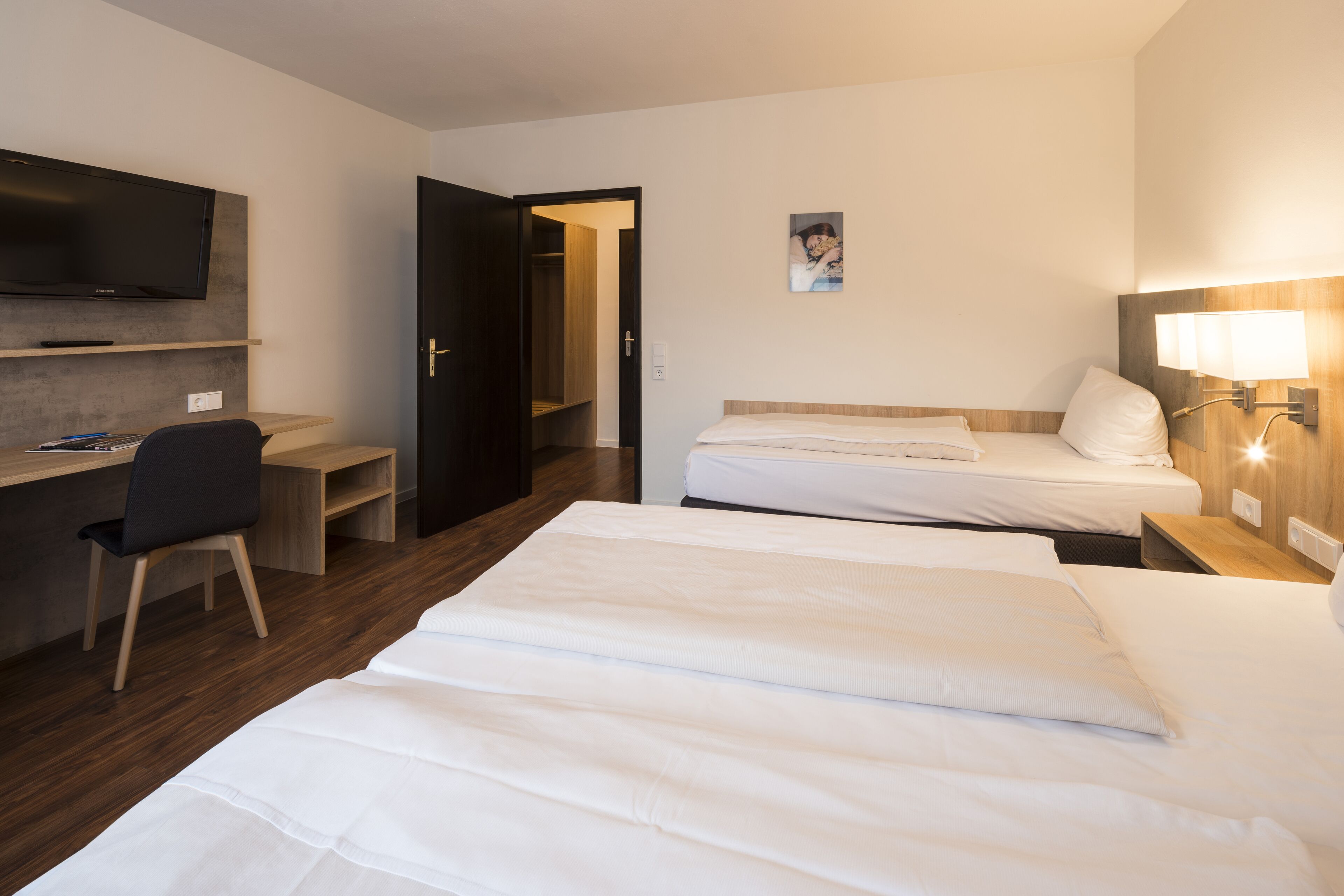 A hotel room with two beds, a desk with a chair, a TV mounted on the wall, and a bedside lamp.