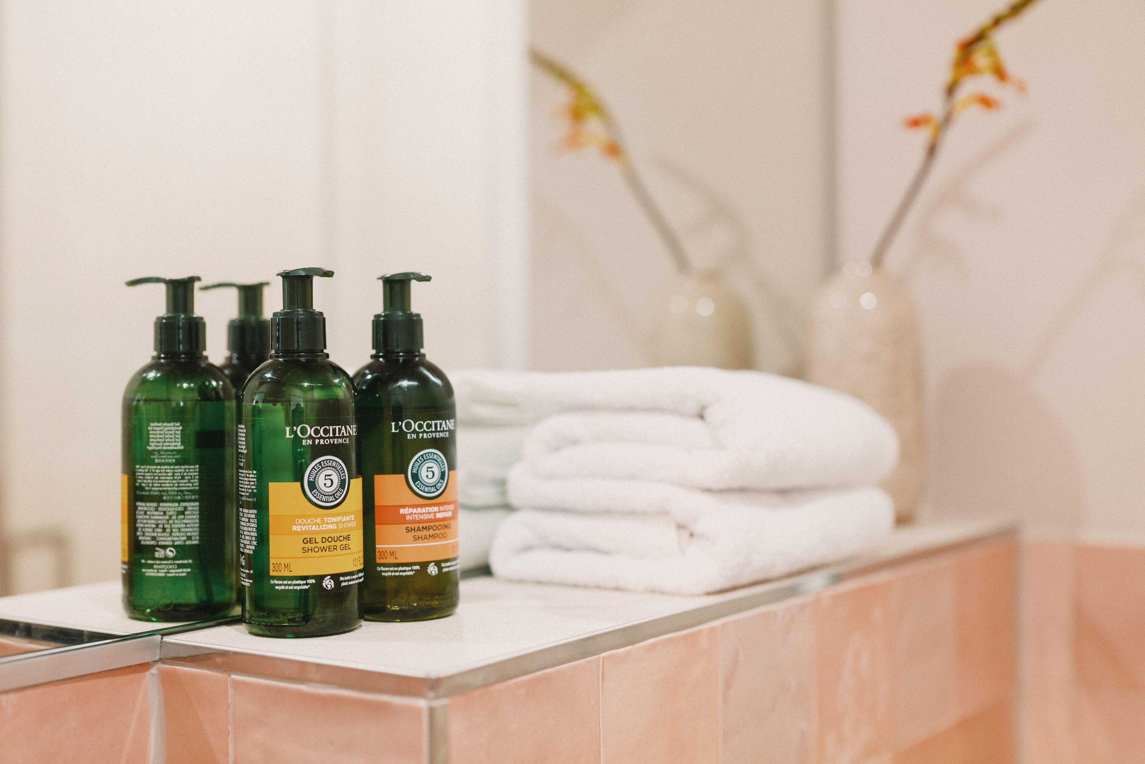Et badeværelsesbord med tre flasker L'OCCITANE shower gel og shampoo, samt en stak hvide håndklæder.