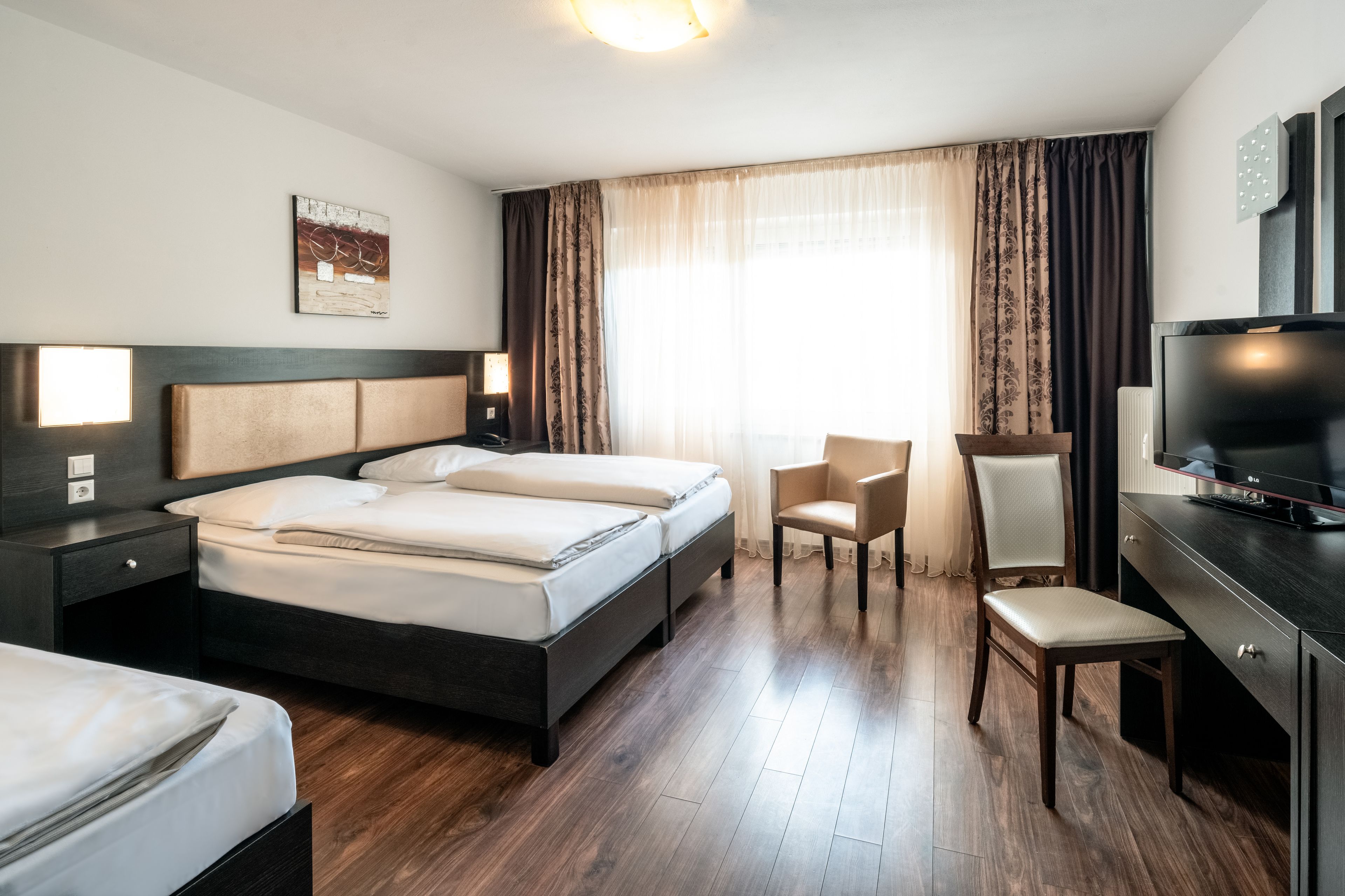 Habitación de hotel con una cama grande, un escritorio con televisor, una silla y un ventanal con cortinas.
