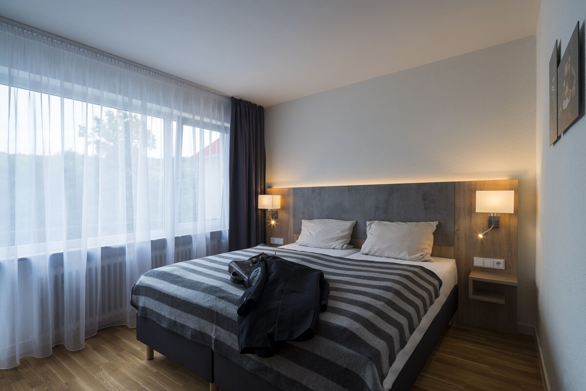 Una habitación de hotel con cama, mesita de noche, lámpara y ventana con cortinas translúcidas.