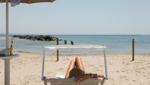 A beach scene with a person lying on a beach chair under a striped umbrella, a bottle, and a pair of sunglasses on a table. - Tonnara di Sciacca (Sciacca)