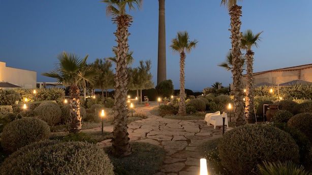 A garden pathway with palm trees, a stone path, and a tall chimney in the background. - Tonnara di Sciacca (Sciacca)