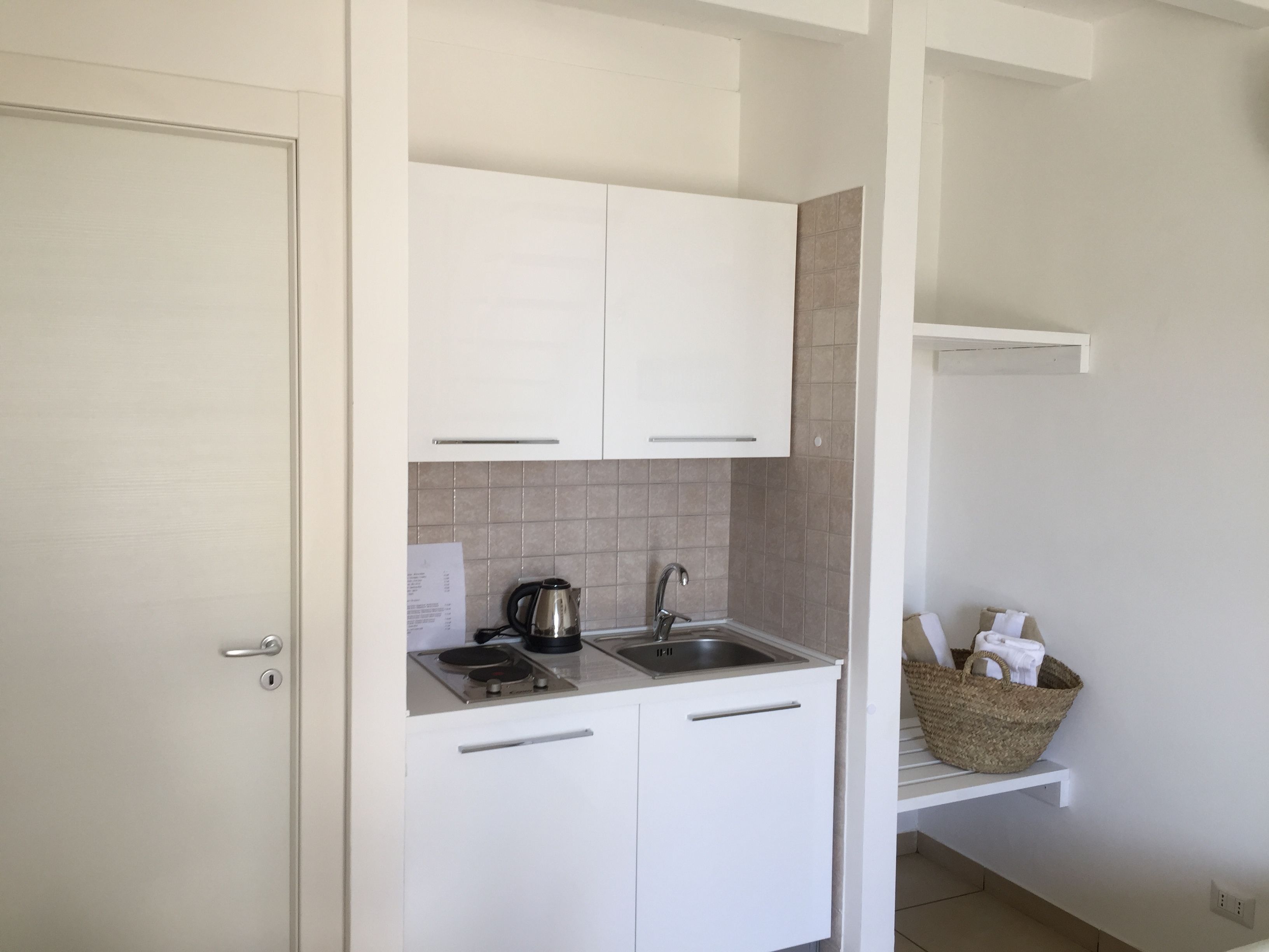Een kleine keuken met witte kastjes, een roestvrijstalen spoelbak en een waterkoker. Een mand met handdoeken staat op een plank.