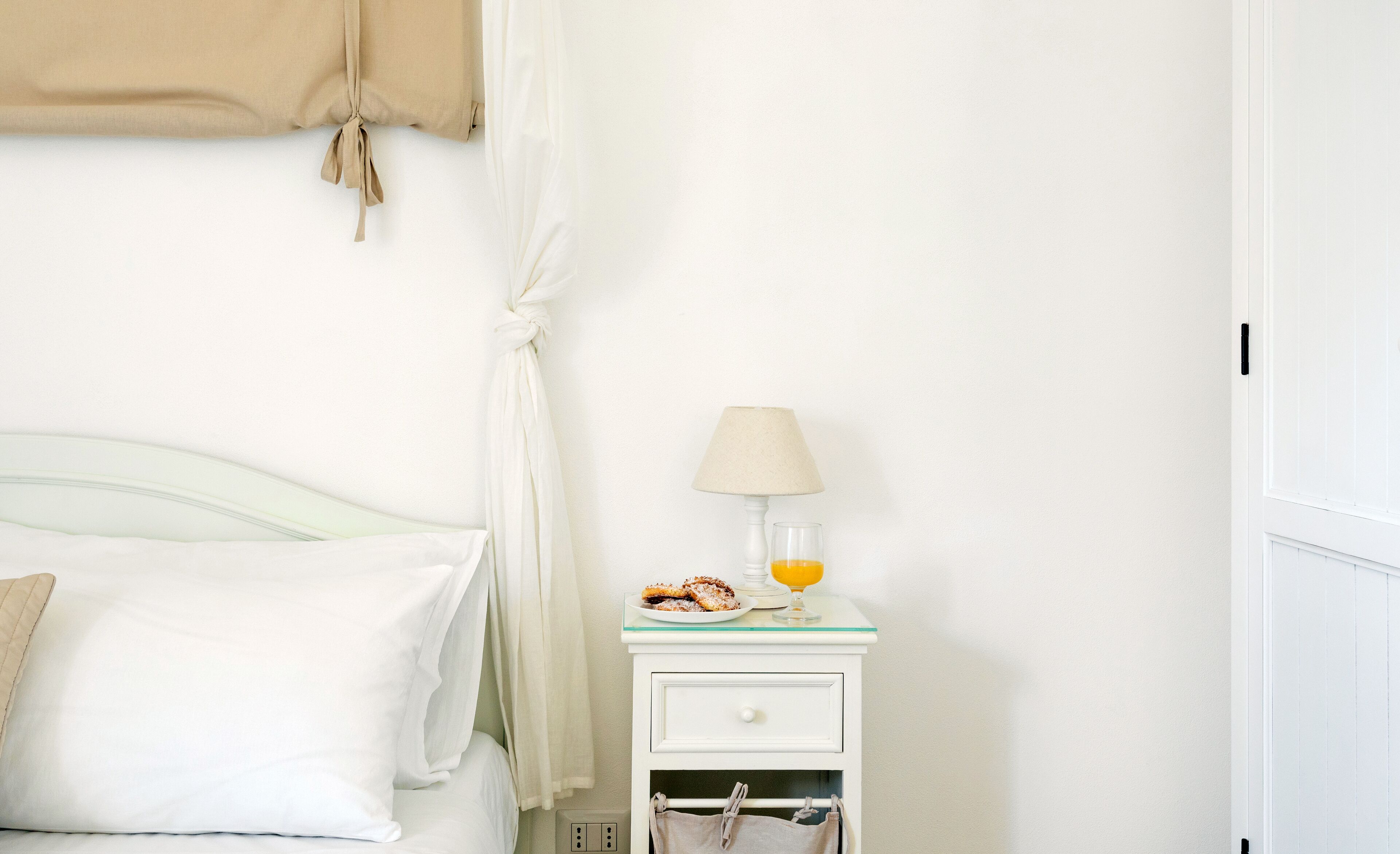 A neatly made bed with white linens and a beige throw blanket. A small bedside table with a lamp and a plate of food is visible.