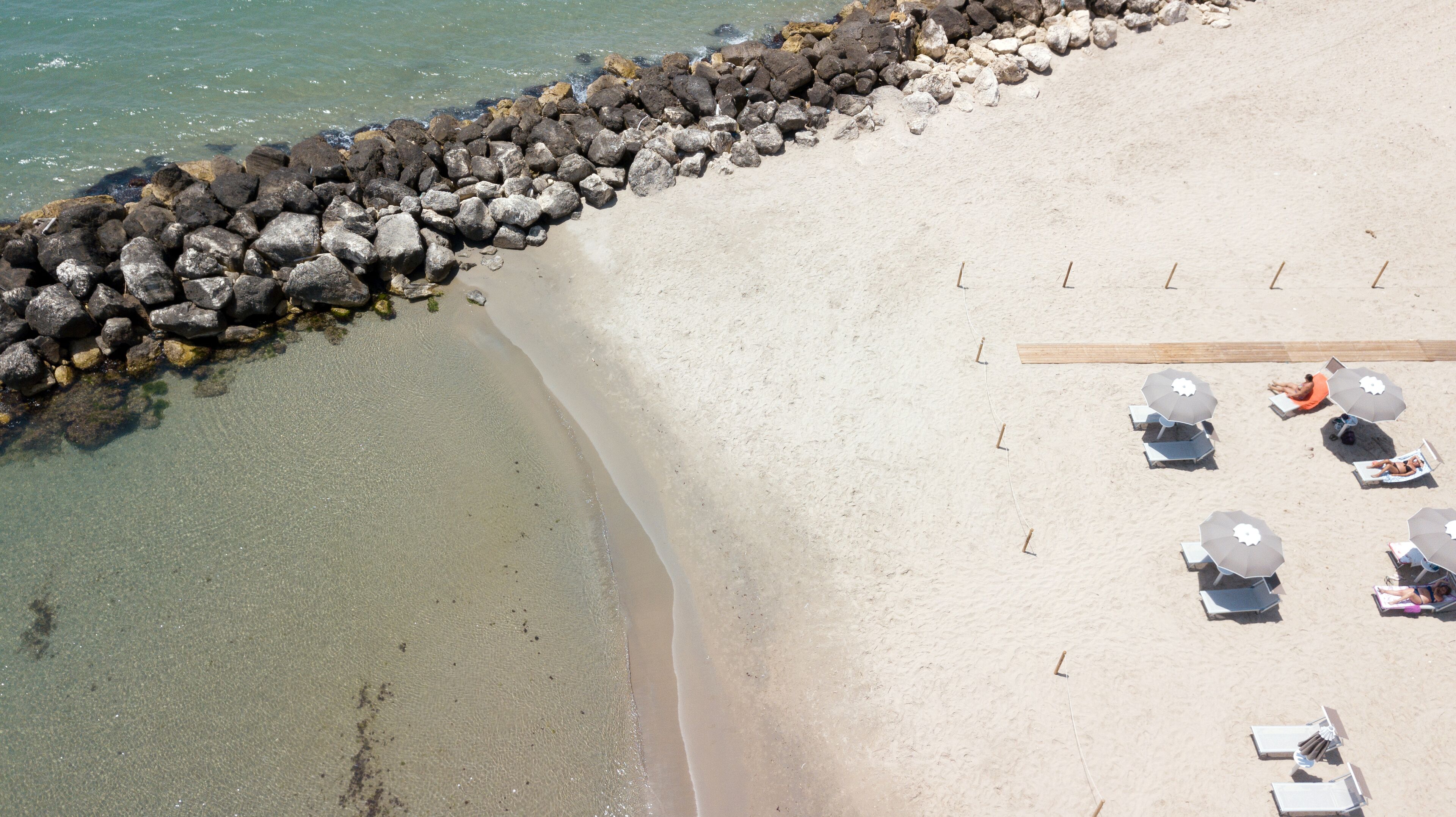 Vista aérea de una playa con un rompeolas rocoso, aguas poco profundas y cristalinas, y varios sombrillas y sillas de playa.