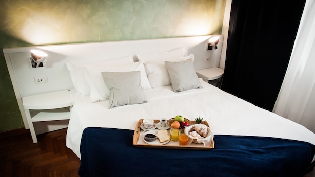 Un lit bien fait, avec du linge de lit blanc, un plateau de petit-déjeuner comprenant des toasts, des fruits et des boissons, et deux lampes de chevet.