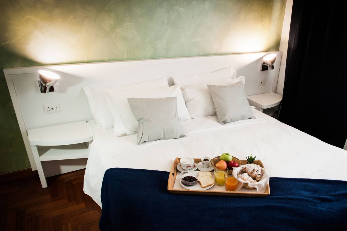 Un lit bien fait, avec du linge de lit blanc, un plateau de petit-déjeuner comprenant des toasts, des fruits et des boissons, et deux lampes de chevet.