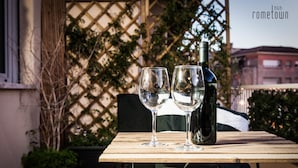 Une table avec deux verres à vin et une bouteille, placée devant un treillage orné de verdure, avec un bâtiment en arrière-plan.
