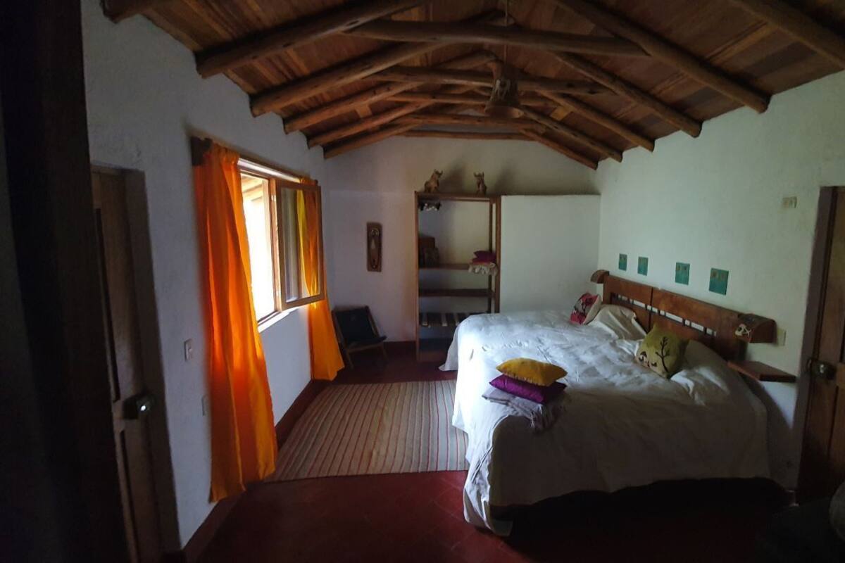 A bedroom with a wooden ceiling, a bed with pillows, a window with curtains, and a wooden door.