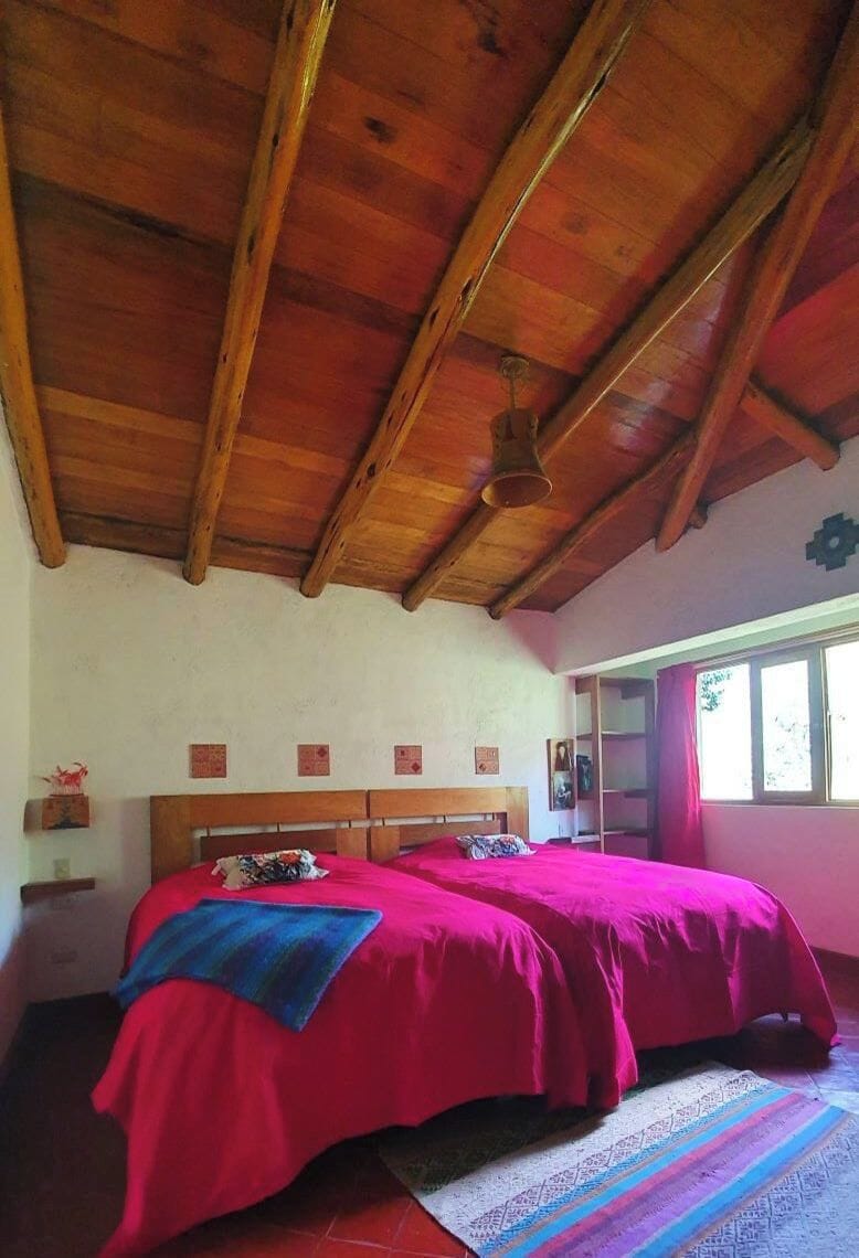 A room with a wooden ceiling, a bed with red bedding, a patterned rug, and a window with curtains.