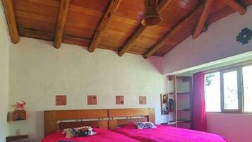 A room with a wooden ceiling, a bed with red bedding, a patterned rug, and a window with curtains.