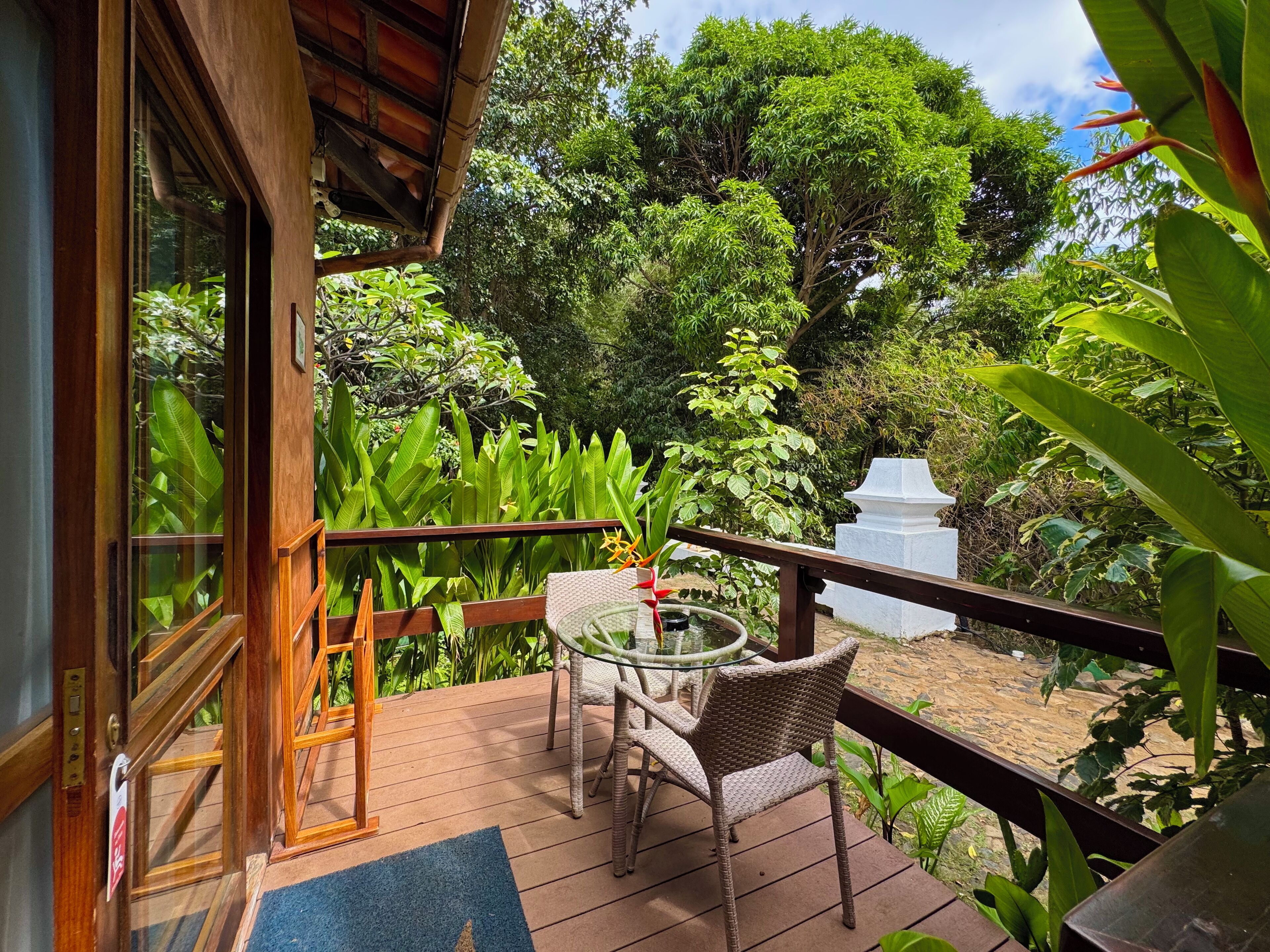 Une terrasse en bois avec une porte vitrée donnant sur une pièce, le tout entouré d’une végétation luxuriante et agrémenté d’un piédestal blanc.