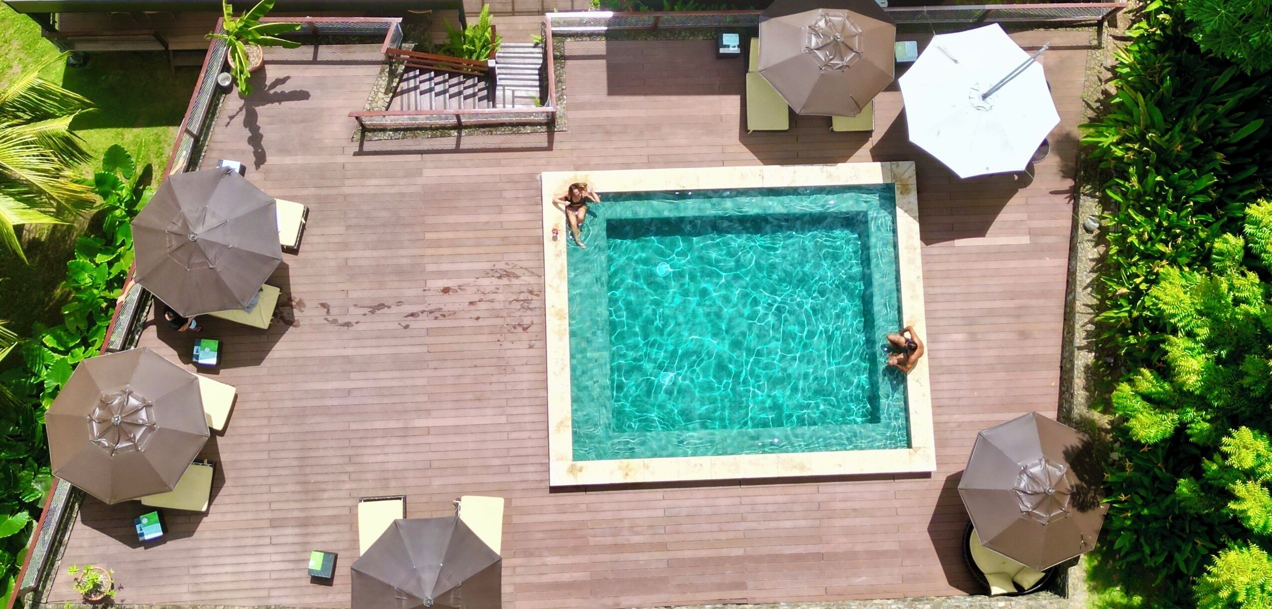 Ein rechteckiger Pool mit einer gefliesten Umrandung, umgeben von einer Holzterrasse. Vier Sonnenschirme und zwei Liegestühle sind vorhanden.