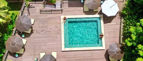 Ein rechteckiger Pool mit einer gefliesten Umrandung, umgeben von einer Holzterrasse. Vier Sonnenschirme und zwei Liegestühle sind vorhanden.