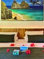 Habitación de hotel con cama, almohadas, un cuadro enmarco de un paisaje costero y flores rojas dispersas.