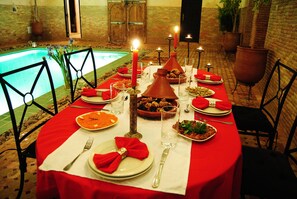 A dining table set for a meal with a pool view, surrounded by chairs and candle-lit tables. - Riad Romance (Marrakech)