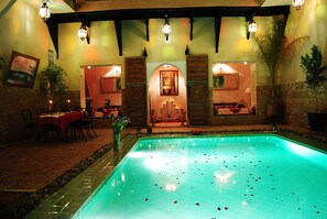 A swimming pool with a tiled edge, surrounded by a brick floor and a low wall. There are tables and chairs on the left, and a decorative archway in the center. - Riad Romance (Marrakech)