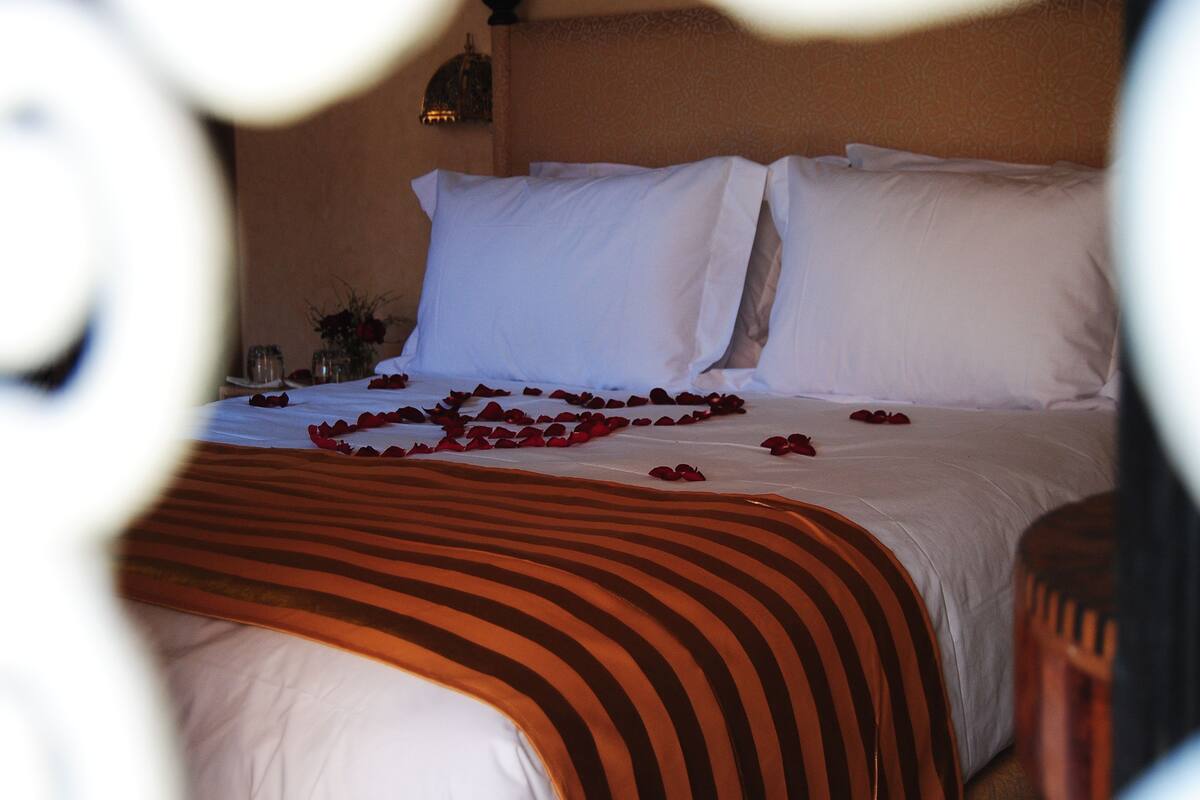 A neatly made bed with white linens and a striped blanket, a vase with flowers, and a small table with a candle and a glass.