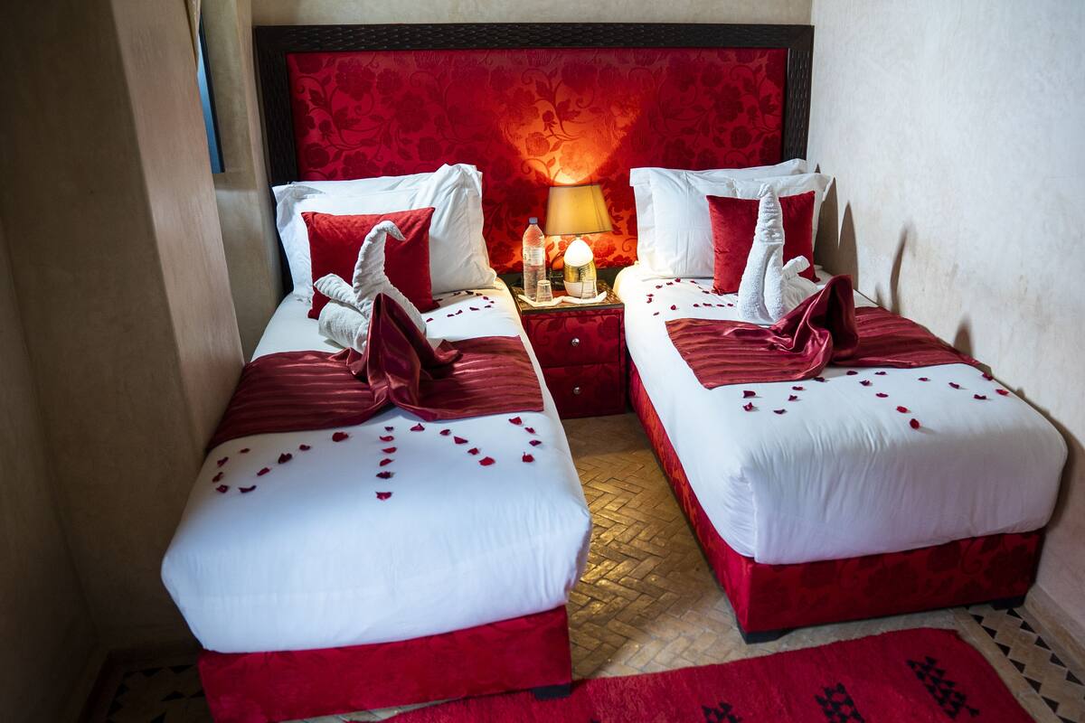A hotel room with two beds, red and white bedding, a red headboard, a nightstand with a lamp, and a red rug.
