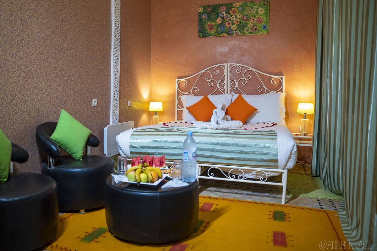 A hotel room with a bed, two chairs, a table with fruits and a bottle, and a painting on the wall.