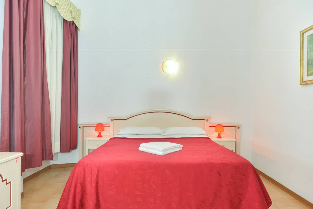 A neatly made bed with a red bedspread, two bedside lamps, a nightstand, and a wall-mounted light fixture.